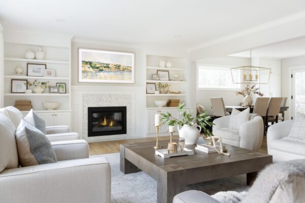 A bright, open living room with a cozy fireplace, custom white built-in shelves, and neutral-toned furniture arranged around a wood coffee table with candles and greenery.