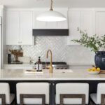 A bright kitchen island with a gold faucet, bowl of lemons, candle, and a large ceramic vase with branches, set against a white herringbone tile backsplash. broomfield kitchen remodeling