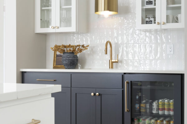 Chic beverage bar with navy lower cabinets, white upper cabinets with glass doors, gold hardware, and a beverage fridge stocked with canned drinks.
