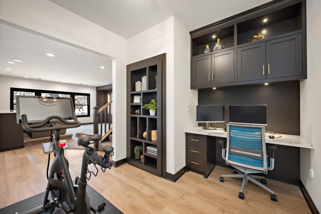 Modern home office with built-in desk and dual monitors adjacent to a Peloton bike on light wood flooring