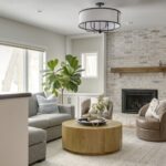 A cozy living room with a light-washed brick fireplace, wooden mantel, built-in cabinets, a gray sectional, a round wooden coffee table, and a fiddle leaf fig tree by the window.