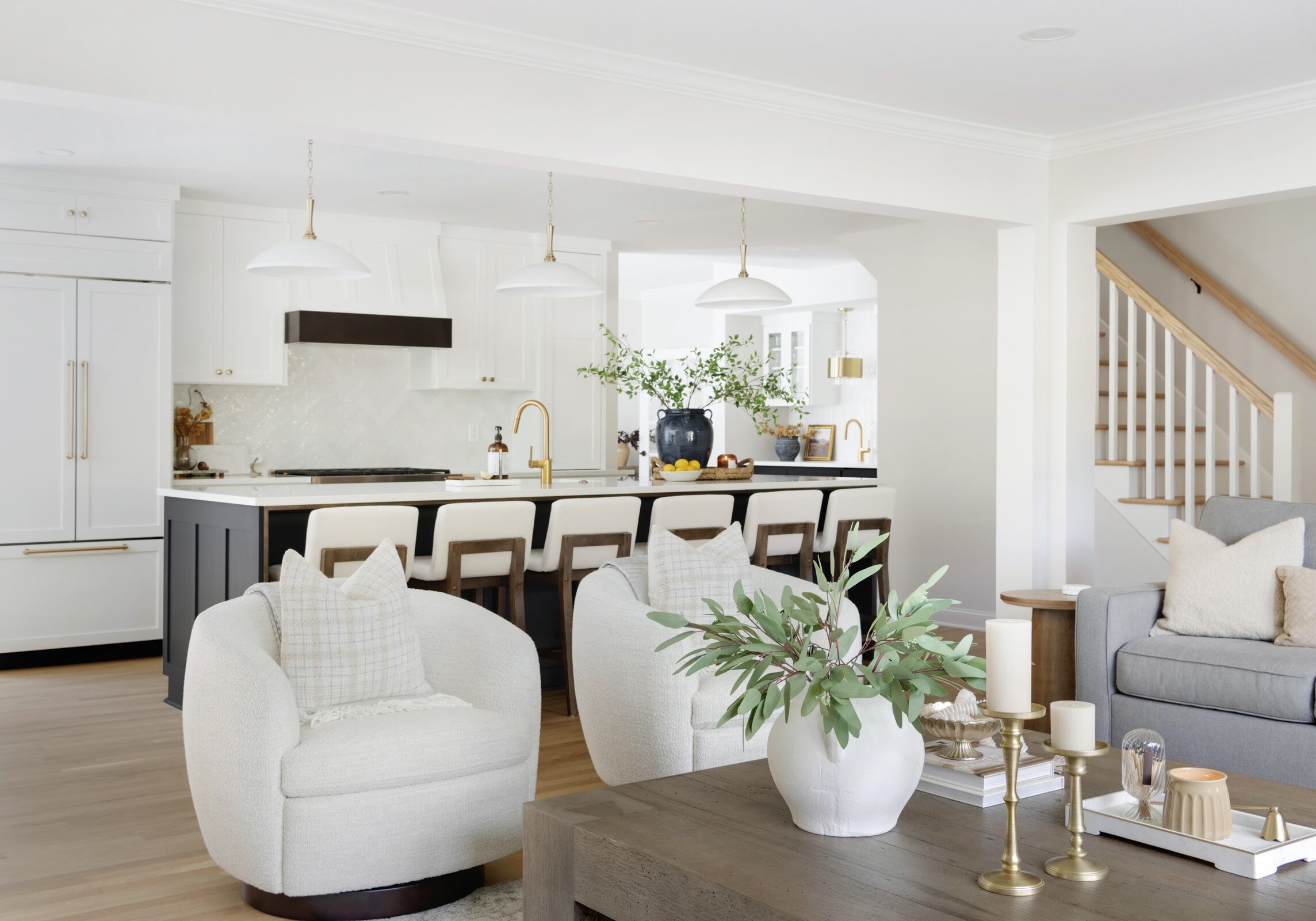 Open-concept living space featuring plush swivel chairs, a rustic coffee table with decor, and a view into a modern kitchen with white cabinetry and gold accents.